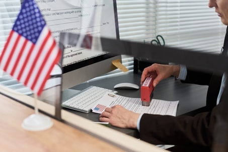 Middle aged Caucasian woman processing visa application at desk, stamping official document with approved mark, working in visa center office with American flag visible on counter